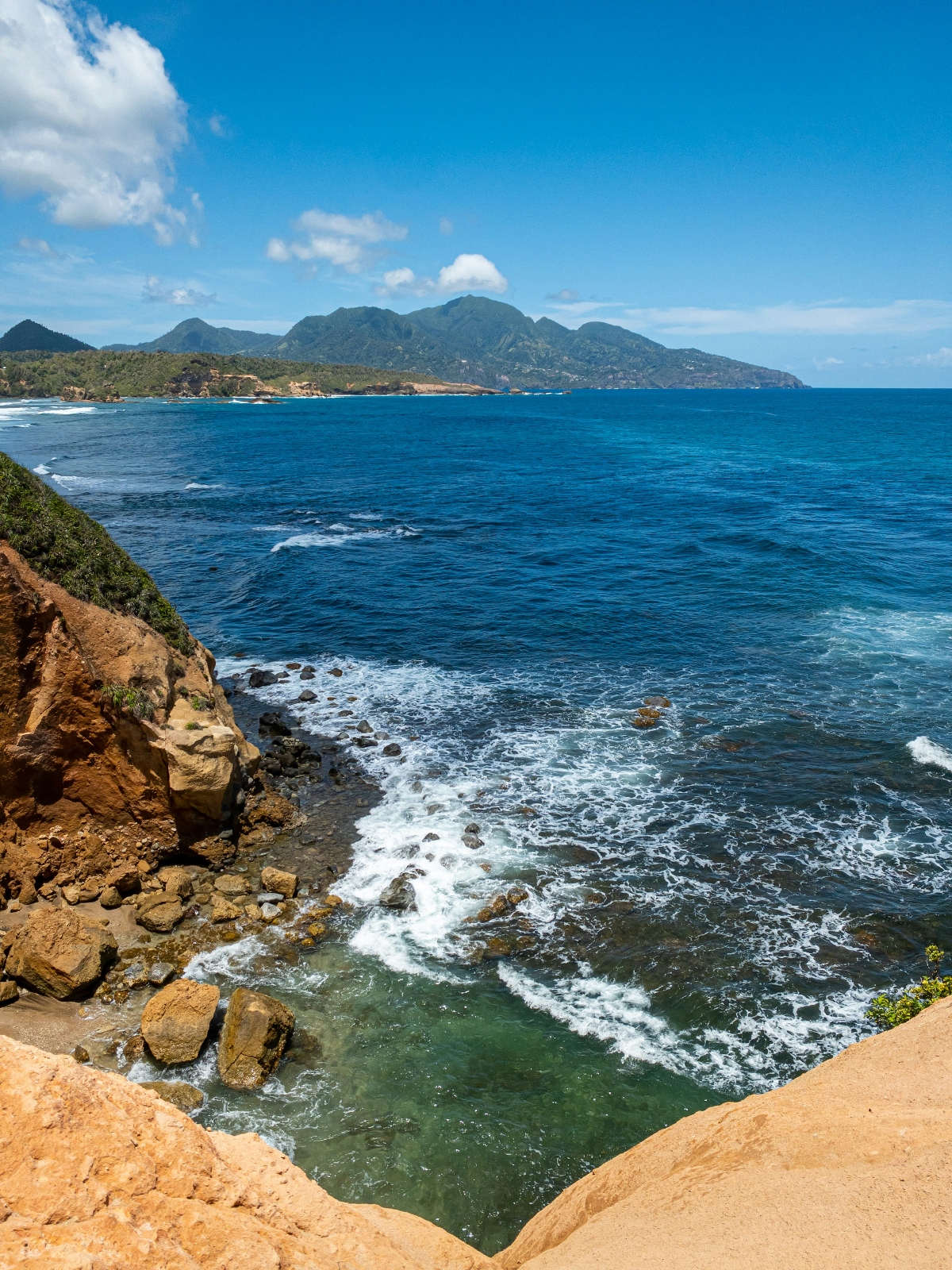 A coastal scene photographed from rocky tan-colored cliffs overlooking deep blue ocean waters with white foam visible along the shoreline and around rocks. A mountainous coastline extends across the background, covered with green vegetation, while several peaks rise against a bright blue sky with scattered white clouds. Waves break against the rocky shore in the foreground, creating patterns of white water in the turquoise-tinted shallows.