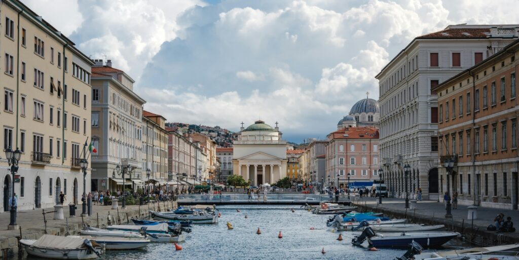 A canal lined with small boats runs through the center of a European coastal town, flanked by multi-story classical buildings in pastel colors. In the background, a neoclassical church or temple with columned portico and dome is centered at the end of the waterway, with hillside buildings visible beyond. Dramatic white cumulus clouds dominate a blue sky above the scene, while pedestrians occupy the waterfront plaza areas on both sides of the canal.