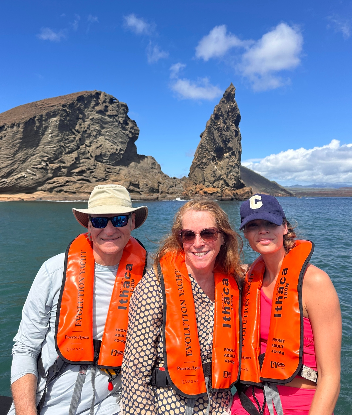 Three people on a family vacation with adult children, wearing bright orange life jackets pose on a boat with a striking rocky formation and blue sky in the background. They are smiling, with the iconic Pinnacle Rock on Bartolomé Island in the Galápagos Islands behind them.
