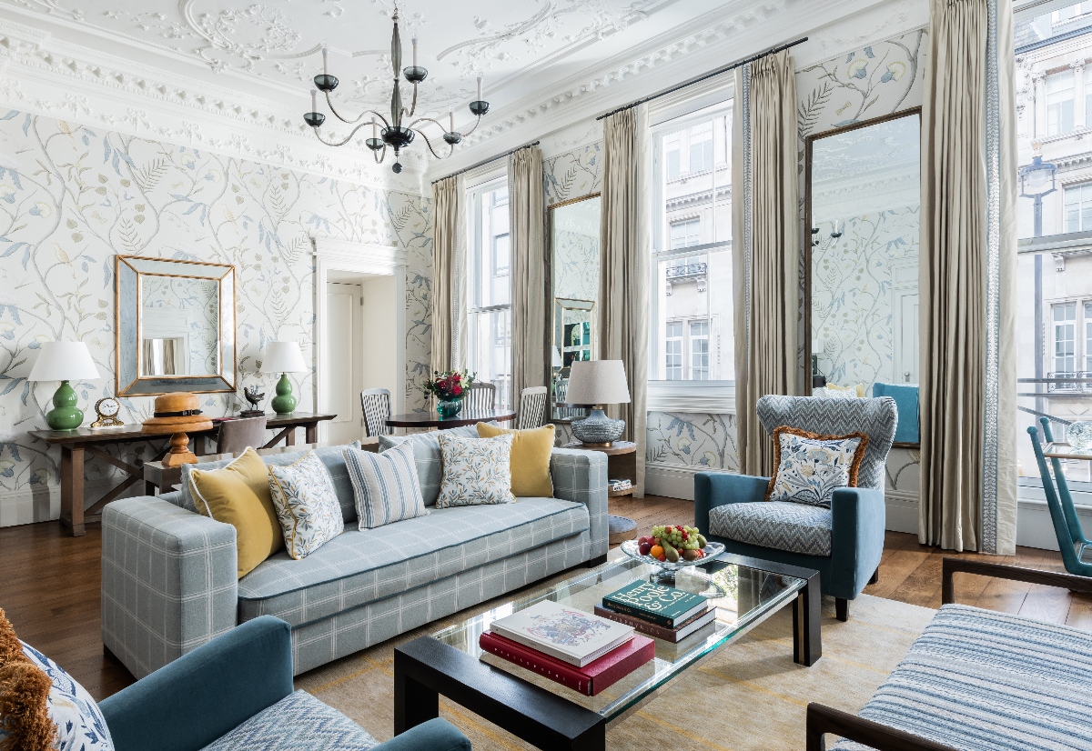 An elegant hotel suite living room features ornate ceiling moldings, delicate blue and white floral wallpaper, and a light blue checkered sofa adorned with yellow and patterned pillows arranged around a glass coffee table. The space showcases classic European luxury with tall windows dressed in cream curtains, traditional blue upholstered chairs, a black wrought iron chandelier, and views of historic London architecture through the windows.