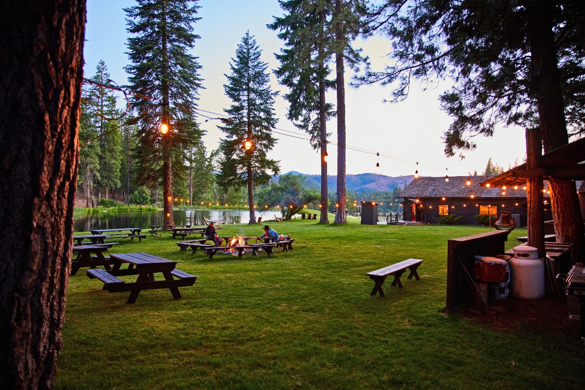 Grassy outdoor area at a lakeside lodge surrounded by tall pine trees, with picnic tables, string lights hanging overhead, and a group of people sitting around a fire pit at dusk. A rustic cabin-style building is lit warmly in the background with forested hills beyond.