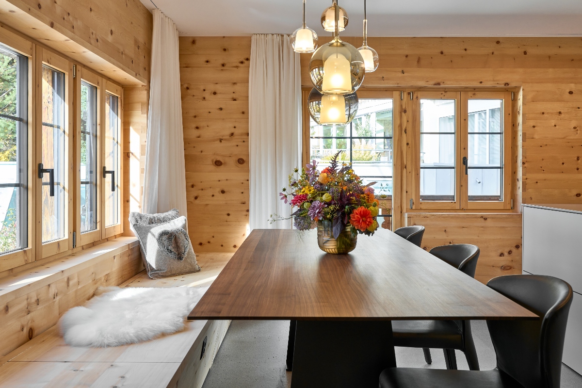 A modern rustic dining room featuring natural wood-paneled walls and a large wooden dining table with black chairs. Above the table hang glass globe pendant lights, while a colorful autumn floral arrangement serves as the centerpiece. Along the windows is a built-in bench with white cushions and pillows, complemented by a white fur rug on the floor. Large windows with white curtains provide abundant natural light, creating a warm, Scandinavian-inspired interior space.