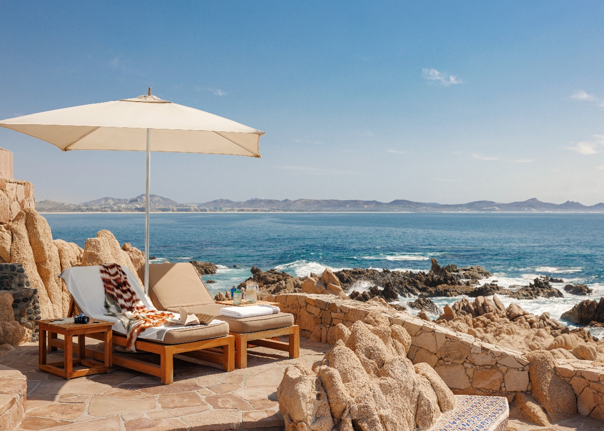A luxury oceanfront terrace features two cushioned lounge chairs with a cream umbrella on a stone patio, overlooking dramatic rocky coastline formations and crashing waves. The scenic vista extends across turquoise blue waters to distant mountainous coastline under a clear sky, creating an idyllic Los Cabos resort setting.