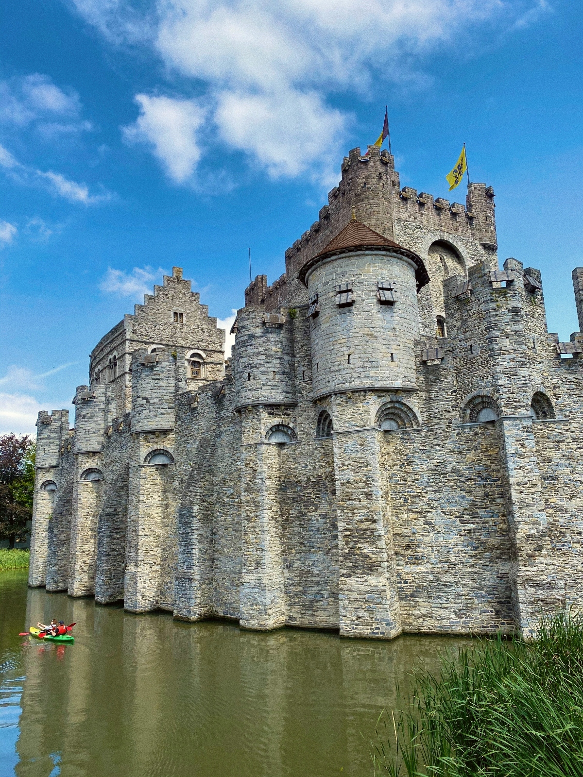A medieval stone castle with multiple cylindrical towers and crenellated walls is surrounded by a water-filled moat, with colorful flags flying from the battlements. A kayaker in red paddles in the moat at the base of the weathered gray stone fortification, which features arched windows and doorways. The castle is photographed against a bright blue sky with white clouds, with reeds growing along the water's edge in the foreground.