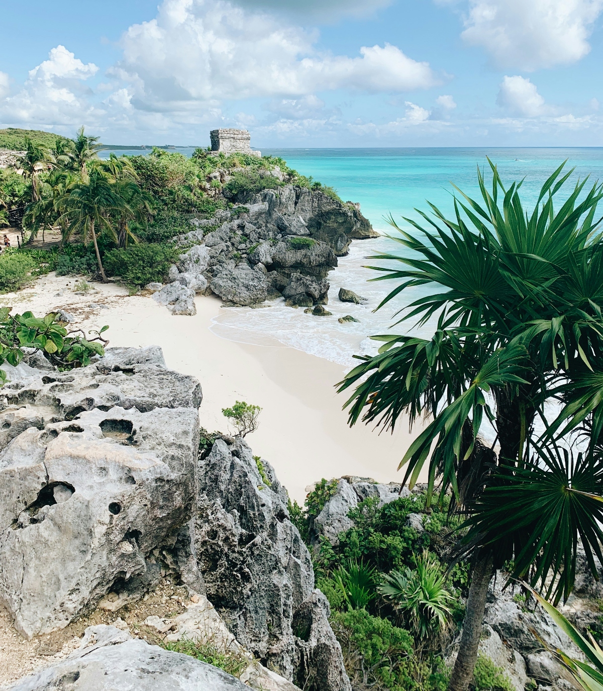 An elevated view of a small sandy beach nestled between weathered limestone cliffs covered with tropical vegetation, including palm trees and green shrubs, with turquoise ocean waters visible beyond. An ancient stone structure sits atop the cliff in the background, while porous gray rock formations frame the foreground under a partly cloudy sky.