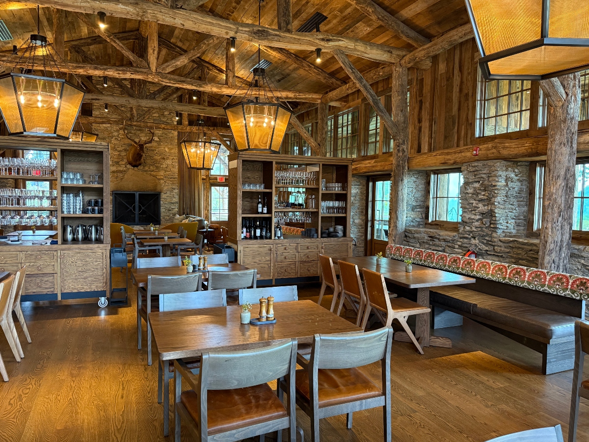 A rustic lodge-style dining room features exposed wooden beam ceilings, stacked stone walls, and large lantern-style pendant lights suspended overhead. The space contains multiple wooden dining tables with chairs, built-in shelving displaying glassware and bottles, and a banquette with patterned orange upholstery. Natural wood support columns and large windows create an open, mountain retreat atmosphere with a mounted deer or elk head visible on the stone wall.