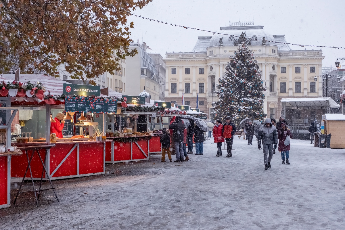 A snowy Christmas market scene with visitors walking among red and white wooden market stalls while snow falls heavily. A large decorated Christmas tree stands in the center of the square, with a grand neoclassical building with cream-colored facade visible in the background. The market area is strung with overhead lights, and visitors bundled in winter clothing browse the stalls along the snow-covered walkway.