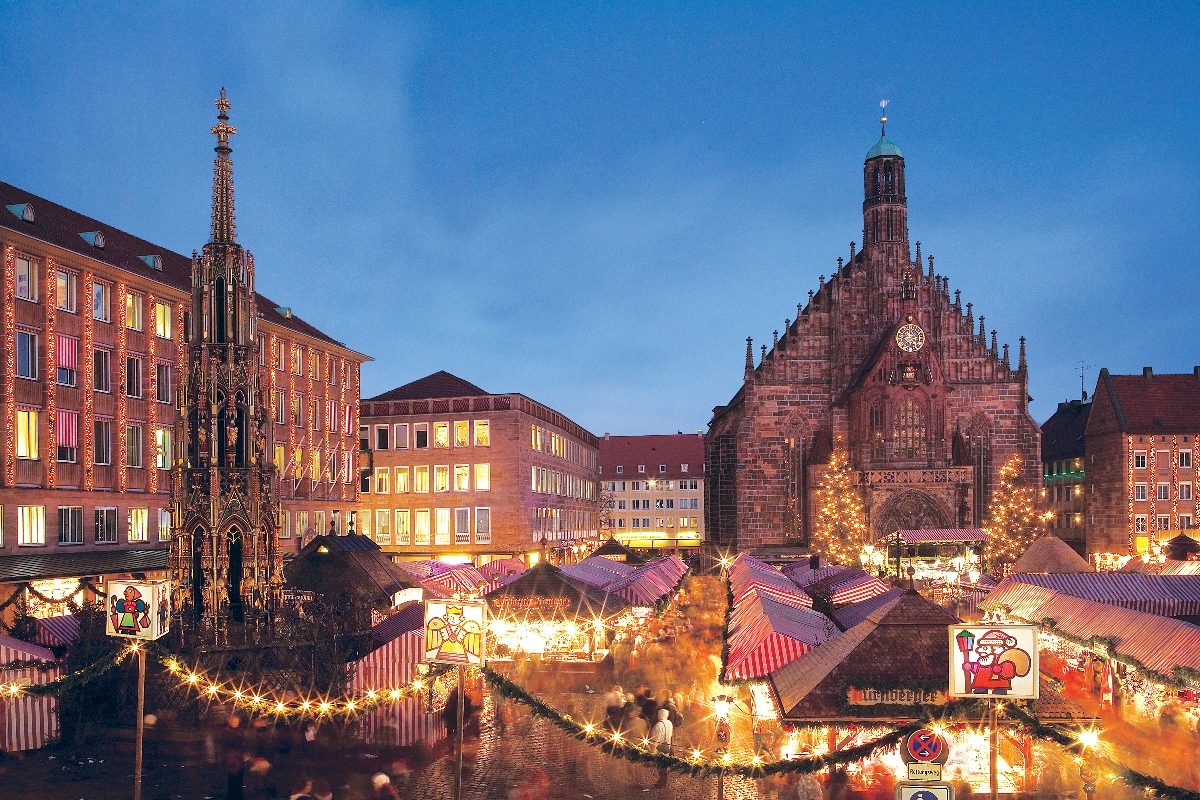 An evening view of Nuremberg's Christmas market in the Hauptmarkt square, showing numerous traditional market stalls with red and white striped awnings illuminated by warm lights. The Gothic Frauenkirche (Church of Our Lady) with its distinctive clock tower dominates the right side of the frame, while the ornate Schöner Brunnen (Beautiful Fountain) spire stands on the left. Crowds of visitors browse the market stalls under a clear blue twilight sky, with decorated Christmas trees and illuminated historic buildings surrounding the square.