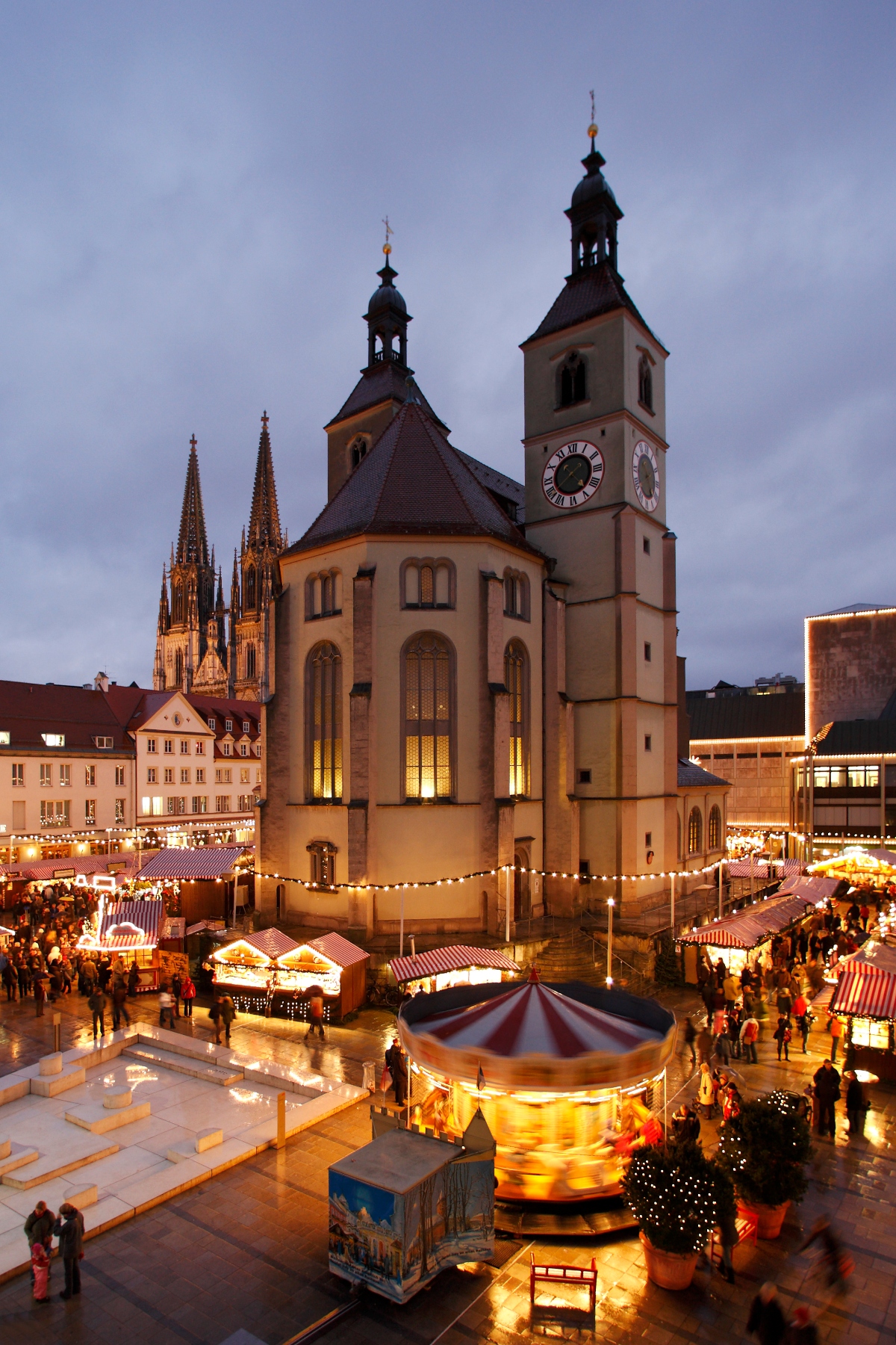An evening view of Regensburg's Christmas market in a town square, dominated by a large illuminated church with twin bell towers topped with distinctive onion domes and featuring clock faces on the right tower. The Gothic spires of Regensburg Cathedral are visible in the background on the left, while the foreground shows market stalls with striped awnings, a brightly lit carousel, and crowds of visitors on the wet plaza. String lights outline the perimeter of the church, and historic buildings frame the square under an overcast twilight sky.