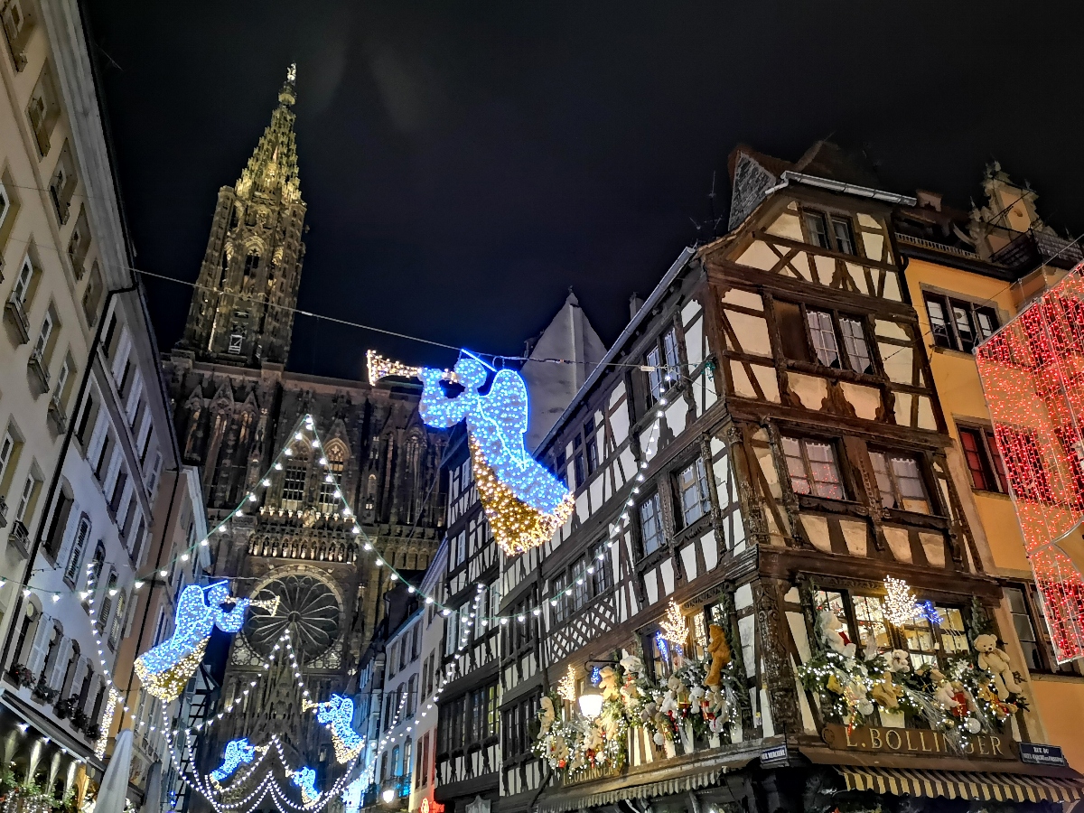 A nighttime view of a narrow street in Strasbourg's historic center, featuring traditional Alsatian half-timbered buildings with white plaster and dark wooden beams, illuminated by Christmas lights and decorations. Large illuminated angel figures made of blue and gold LED lights are suspended between the buildings, with the ornate Gothic spire of Strasbourg Cathedral visible in the background against a dark night sky. The street is adorned with string lights, garlands, and festive decorations, creating a Christmas market atmosphere in the medieval quarter.