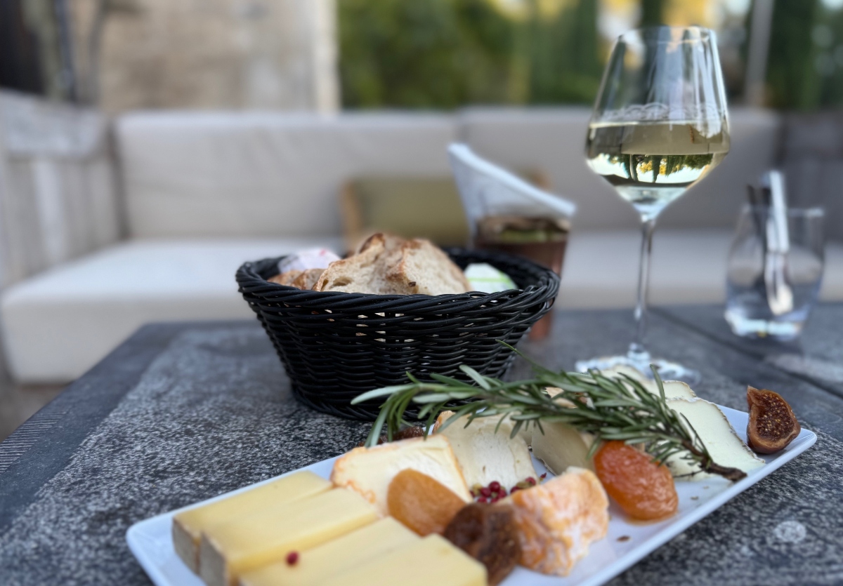 A cheese platter on a white rectangular plate displays several varieties of cheese garnished with fresh rosemary, dried fruits, and what appears to be nuts, served on a dark textured surface. A black woven basket filled with sliced bread sits beside the plate, accompanied by a glass of white wine and a water glass. The setting appears to be an outdoor dining area with soft-focused furnishings visible in the background.