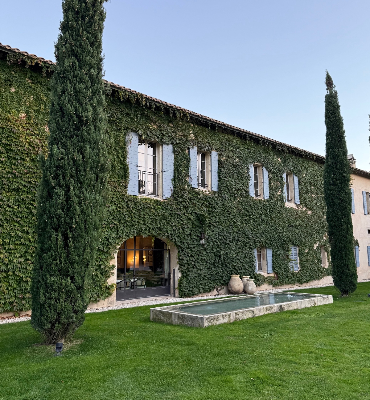 A two-story stone building with a terracotta tile roof is almost entirely covered in green ivy, with white-shuttered windows visible on both levels. Tall cypress trees frame the structure on either side, and a rectangular stone water feature sits on a manicured lawn in the foreground. An arched entrance at ground level reveals a glimpse of an interior space.