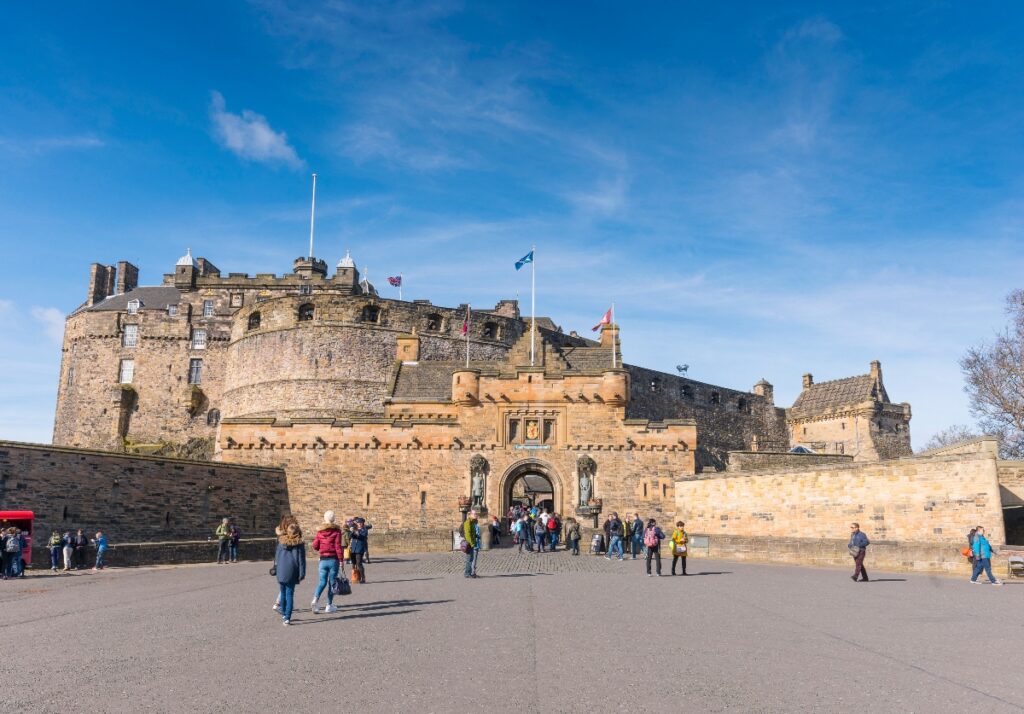 This image shows a large medieval stone castle with multiple towers, battlements, and defensive walls, with several flags flying from flagpoles on the structure. Numerous tourists and visitors are gathered in the paved courtyard area in front of the main entrance gate, some walking around and others standing in small groups. The scene is captured under a bright blue sky with scattered white clouds, highlighting the golden-colored sandstone construction of the historic fortress.