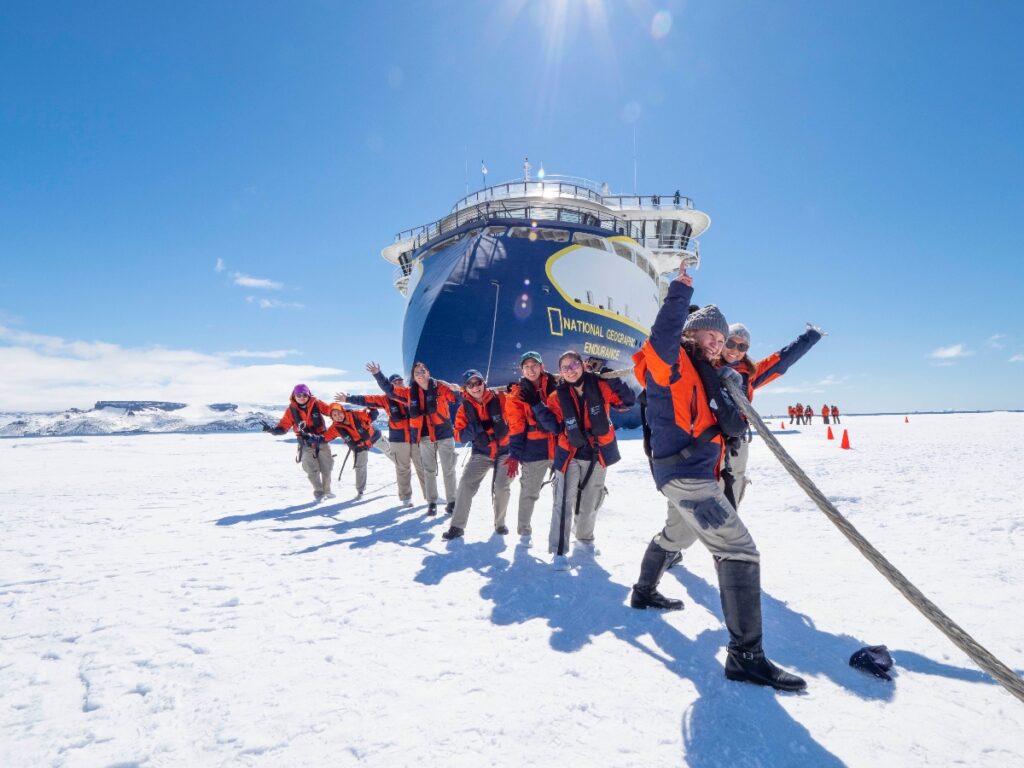 A group of approximately ten people wearing bright orange and black winter expedition gear stand on a snowy landscape with their arms raised in celebration. Behind them is a large blue and white expedition ship with "National Geographic" visible on its hull, positioned on what appears to be frozen terrain. The scene is set against a clear blue sky with distant snow-covered hills visible on the horizon, suggesting a polar or Arctic environment.