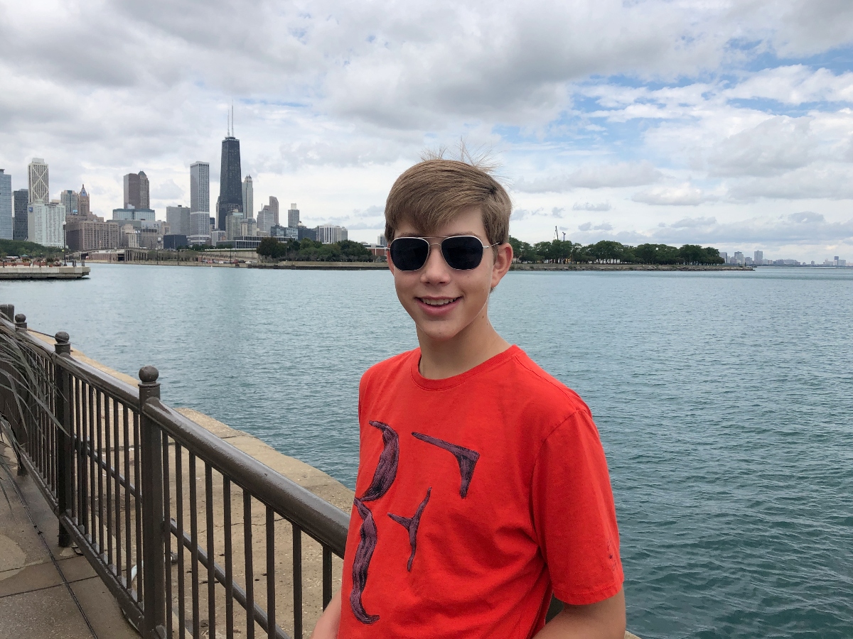 A young person wearing sunglasses and a red t-shirt stands smiling at a waterfront railing with a large body of water behind them. The background features a city skyline with several tall buildings and skyscrapers under a partly cloudy sky. The scene appears to be taken from a lakefront or riverside walkway with a metal railing visible in the foreground.