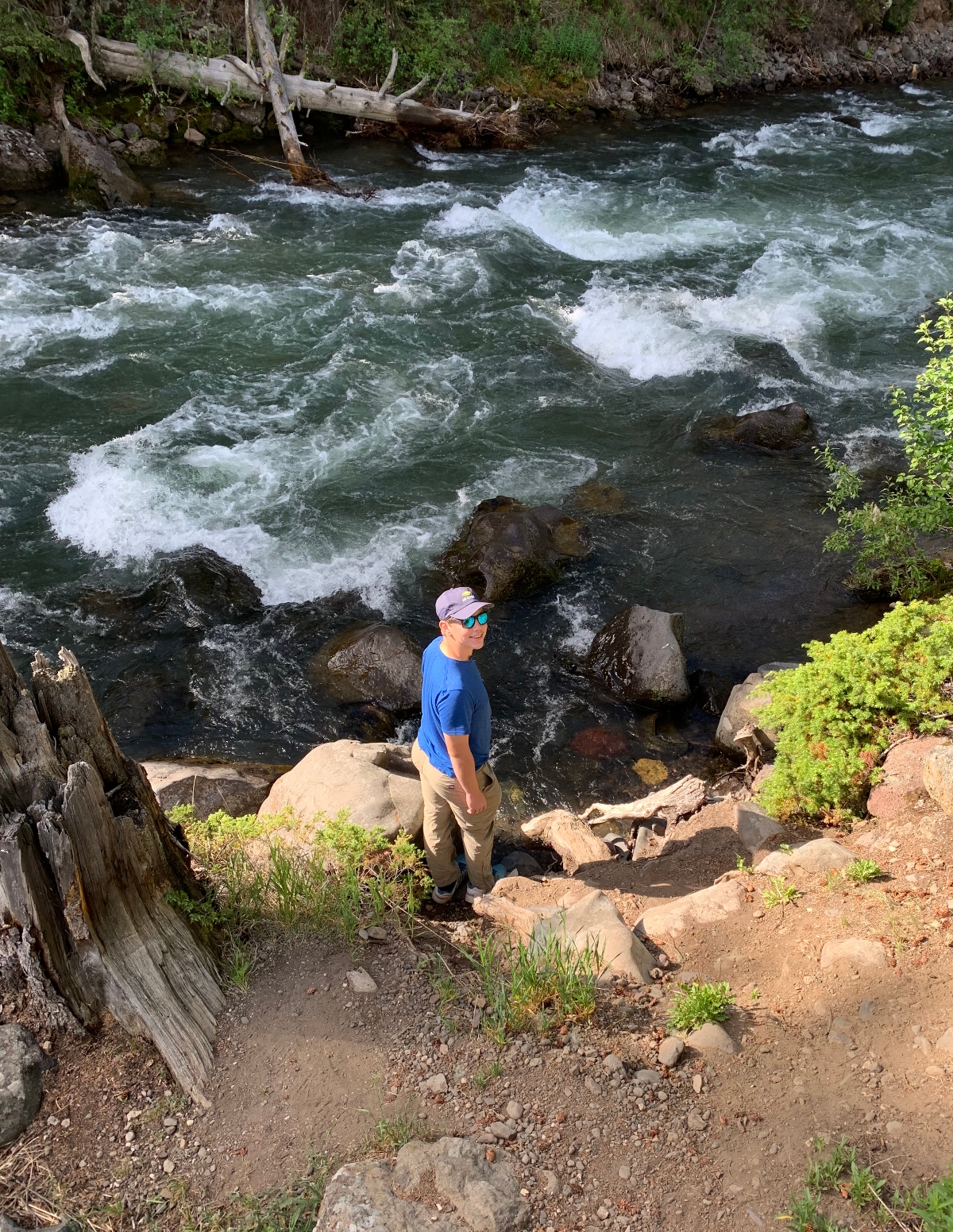 A person wearing a blue shirt, purple cap, and sunglasses stands on a rocky, dirt path beside a fast-flowing river with white water rapids. The scene shows a natural waterway surrounded by large boulders, fallen logs, and lush green vegetation along the banks.