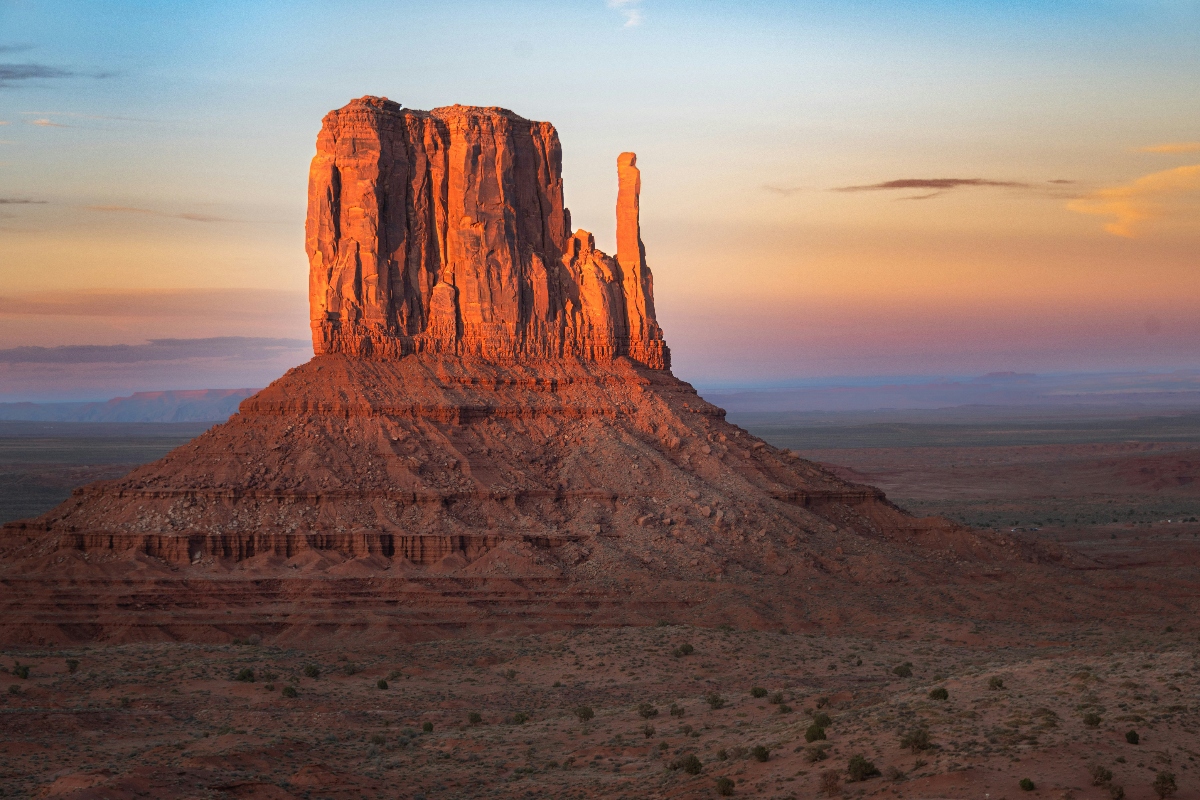 A large sandstone butte with distinctive vertical cliff faces dominates the frame, glowing orange-red in warm sunlight against a partly cloudy sky with sunset or sunrise colors. The massive rock formation rises from a desert landscape with sparse vegetation and rolling terrain extending to distant hills. The lighting creates dramatic shadows and highlights on the layered rock face, emphasizing the rugged geological features of this arid environment.