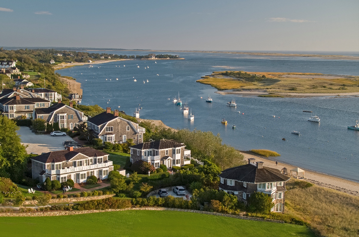 An aerial view shows a collection of traditional shingled houses positioned along a curved coastline, with manicured green lawns extending toward sandy beaches and a protected harbor. The calm blue waters are dotted with numerous sailboats and small vessels at anchor, while low-lying islands and tidal marshlands are visible in the distance. The coastal community appears nestled between the water and inland vegetation under a partly cloudy sky.