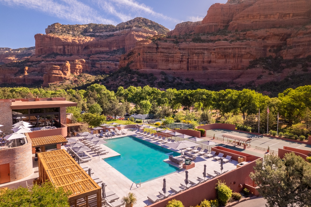 A luxury resort complex features a large rectangular swimming pool surrounded by lounge chairs and umbrellas, with modern southwestern-style buildings constructed in earthy terracotta tones. The property is set against a dramatic backdrop of towering red sandstone canyon walls and rock formations, with lush green trees providing contrast to the desert landscape. The scene is captured under a partly cloudy blue sky, showcasing the integration of contemporary architecture with the natural desert environment.