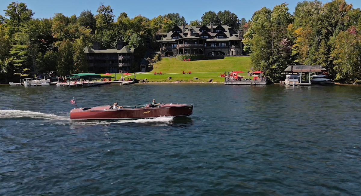 A classic wooden motorboat with passengers cruises across a lake, creating a wake as it moves through the dark blue water. In the background, a large multi-story lodge or mansion with dark wood construction and multiple gabled rooflines sits on a manicured lawn that slopes down to the shoreline. The property features several boat docks along the water's edge and is surrounded by dense forest, with red umbrellas or seating areas visible on the green lawn.
