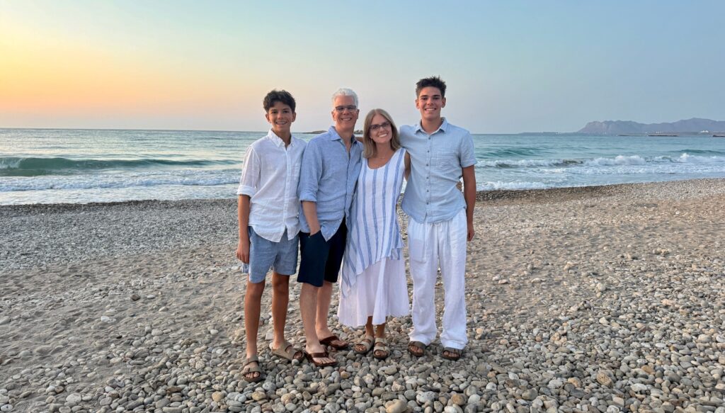 Four people stand together on a pebbled beach wearing coordinated light blue and white clothing, with ocean waves and a sunset sky visible behind them. The beach setting features a rocky shoreline with mountains visible in the distance across the water.
