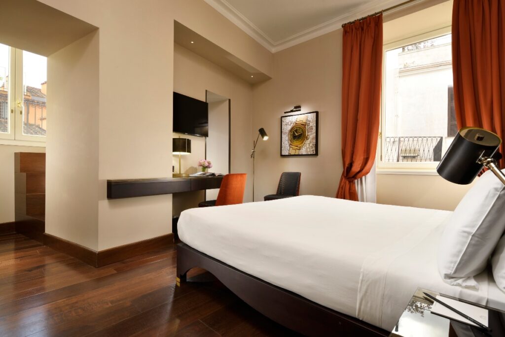 A modern hotel room features a bed with white linens, dark wooden floors, and a built-in desk area with an orange chair positioned near windows with orange curtains. The room has cream-colored walls, a wall-mounted television, and opens to a balcony overlooking neighboring buildings with traditional architecture.