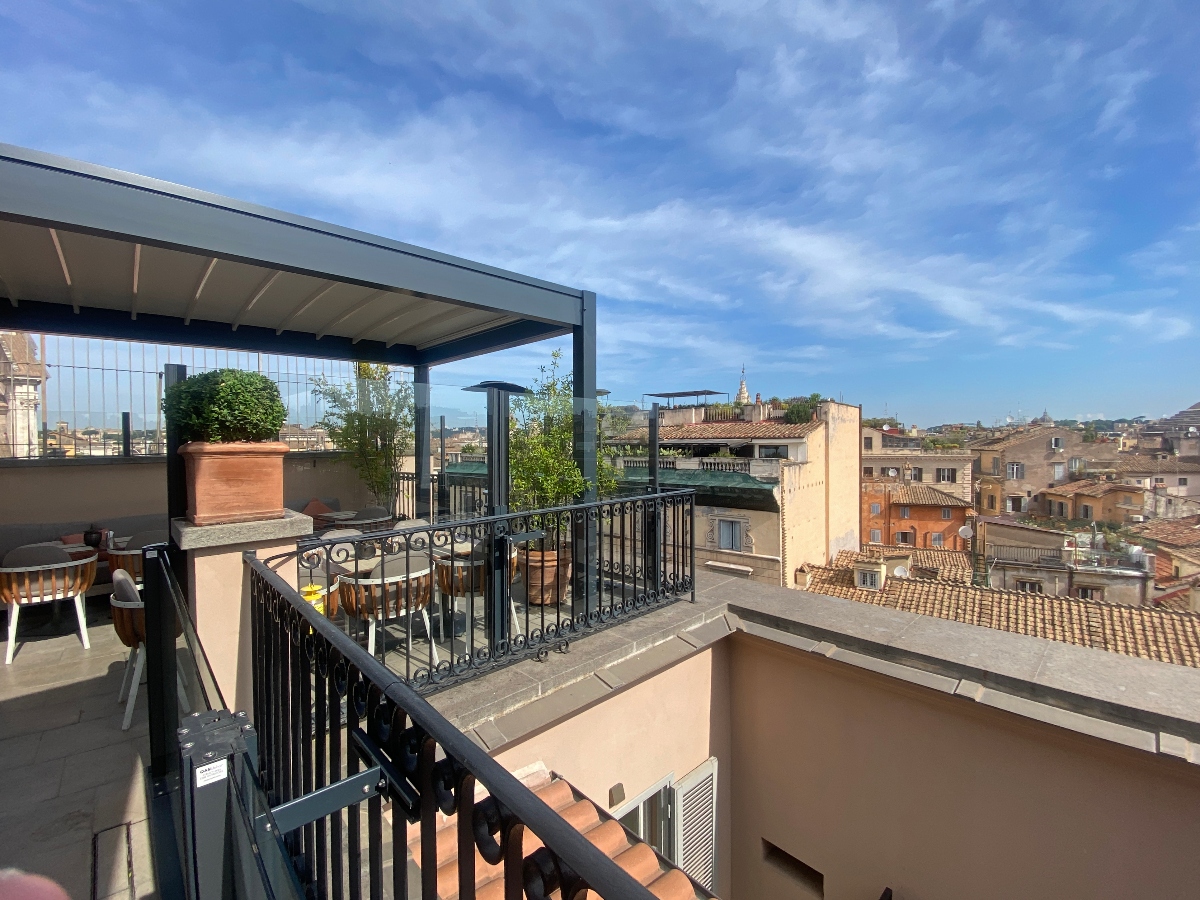 A rooftop terrace features a modern metal pergola with outdoor seating and planters, surrounded by black metal railings. The terrace overlooks a cityscape of historic buildings with terracotta tile roofs under a blue sky with white clouds.