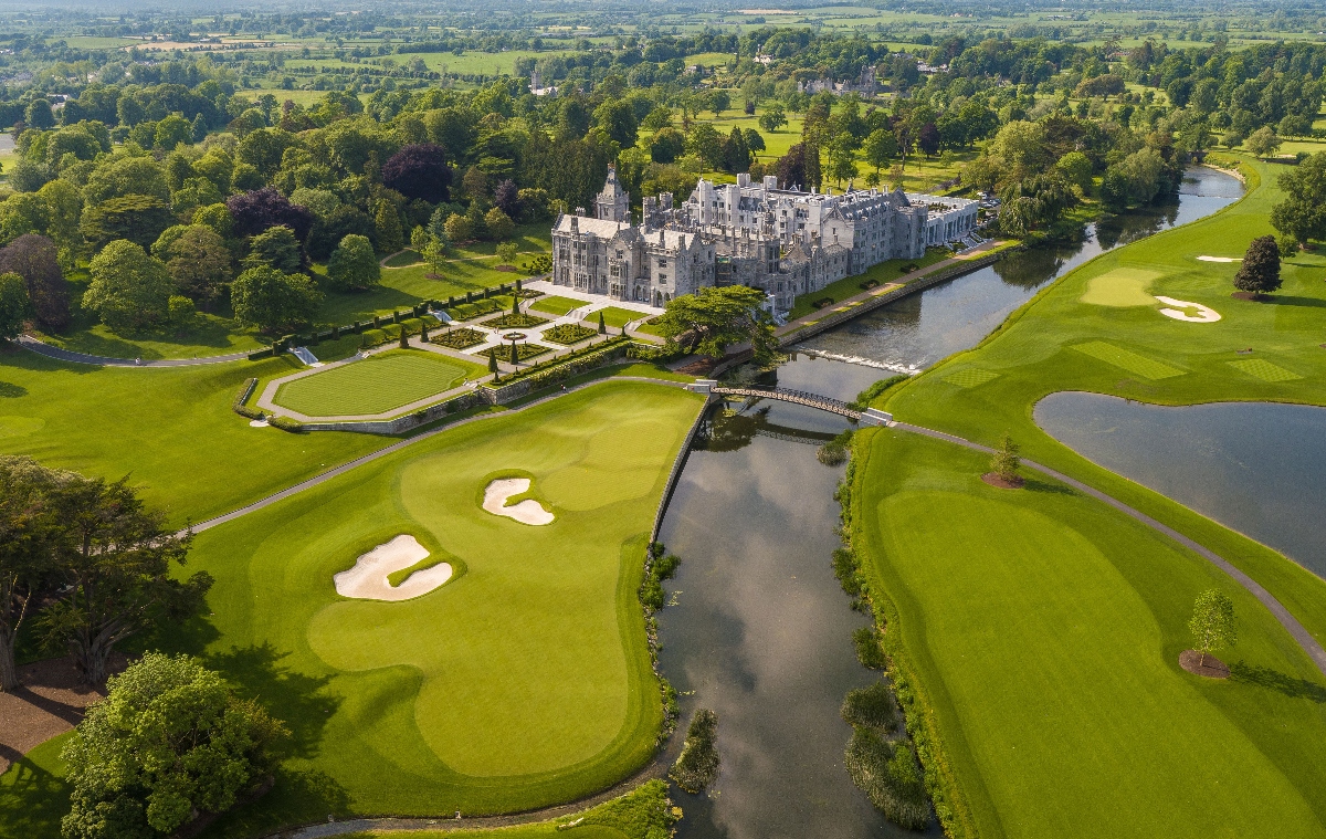 An aerial view shows a large stone castle or manor house with towers and formal architectural features, surrounded by manicured formal gardens with geometric hedge patterns. The property is situated alongside a championship golf course featuring multiple holes with sand bunkers, water hazards, and lush green fairways that wind through the landscape. A river curves through the scene, while expansive countryside with fields, trees, and rolling terrain extends to the horizon under an overcast sky.