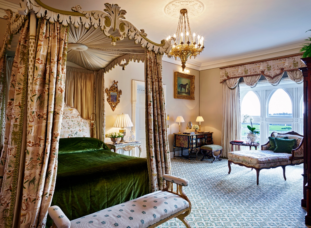 An opulent bedroom features a four-poster bed with an elaborate carved canopy, floral chintz curtains, and emerald green velvet bedding. The room is decorated in a traditional English country house style with floral wallpaper, a crystal chandelier, period furniture including a chaise lounge, and large arched windows with ornate drapery. Antique furnishings, table lamps, and decorative artwork complete the luxurious interior on patterned carpeting.