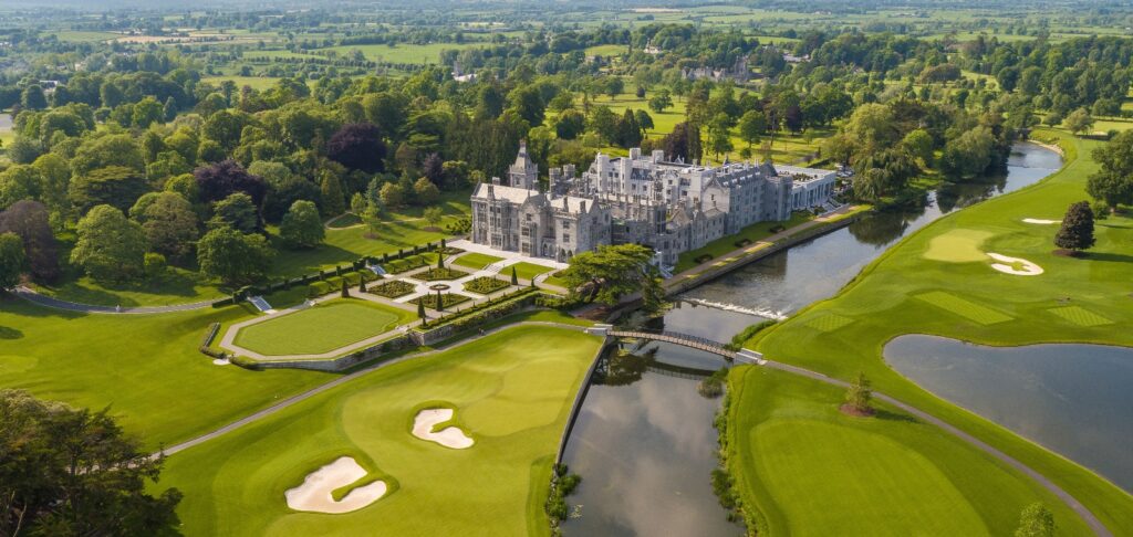 An aerial view shows a large stone castle or manor house with towers and formal architectural features, surrounded by manicured formal gardens with geometric hedge patterns. The property is situated alongside a championship golf course featuring multiple holes with sand bunkers, water hazards, and lush green fairways that wind through the landscape. A river curves through the scene, while expansive countryside with fields, trees, and rolling terrain extends to the horizon under an overcast sky.