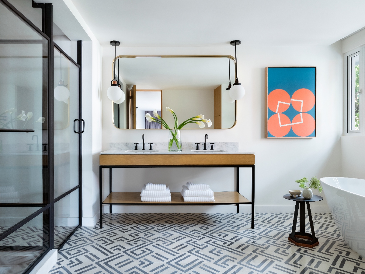 A modern bathroom featuring a floating double vanity with a white marble countertop, wood drawer front, and black metal frame supporting an open shelf with stacked white towels. Above the vanity hangs a large brass-framed mirror with two globe pendant lights, while a glass-enclosed shower with black framing is visible on the left. The space includes geometric patterned floor tiles in gray and white, a freestanding white bathtub on the right, and a geometric abstract art piece in coral and blue tones on the wall.