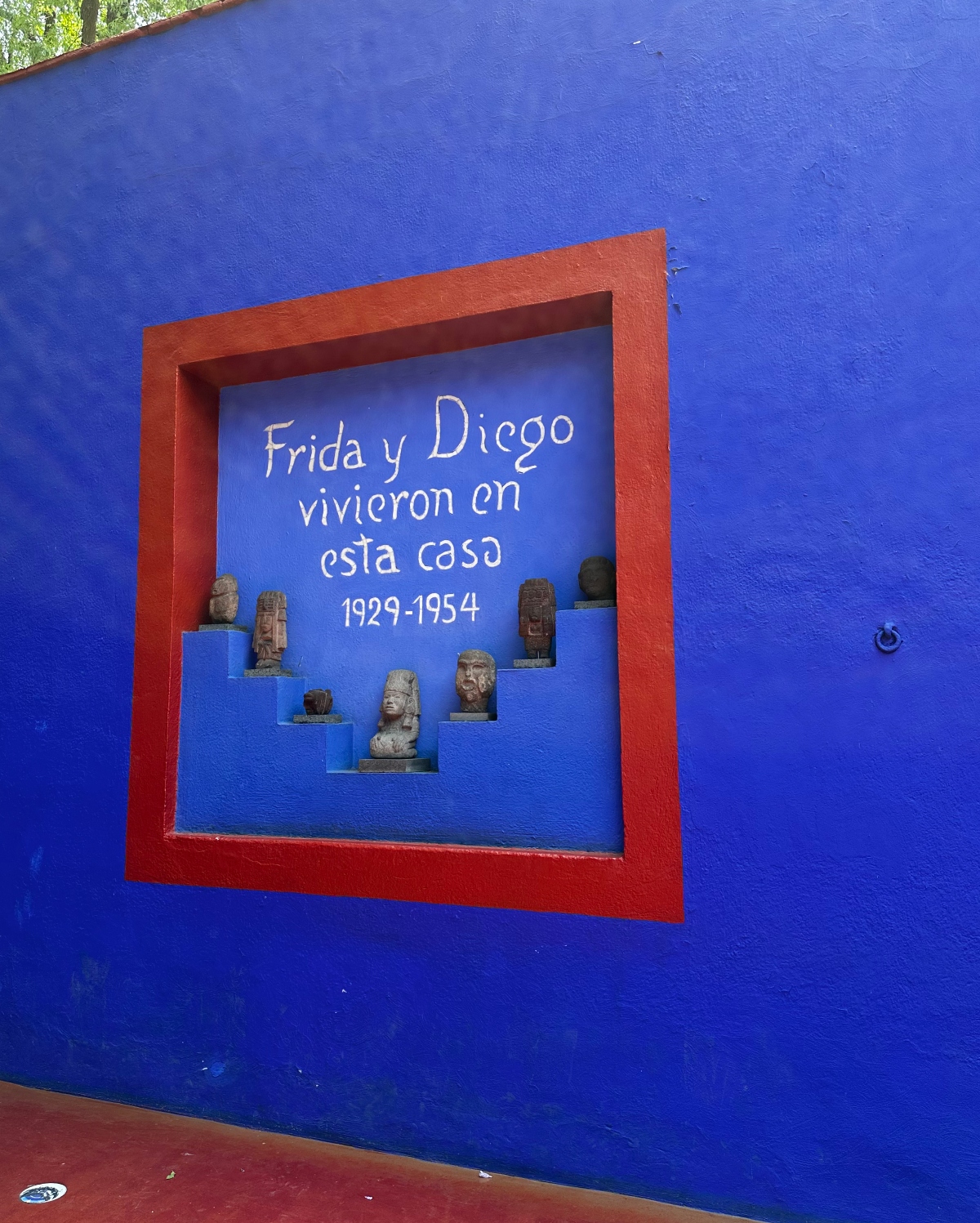 A vibrant cobalt blue wall features a red-framed recessed niche containing white Spanish text that reads "Frida y Diego vivieron en esta casa 1929-1954" (Frida and Diego lived in this house 1929-1954). Small pre-Columbian style sculptural busts and figurines are displayed on stepped blue shelves within the alcove. The wall shows a textured plaster finish with red trim visible at the top and bottom edges of the frame.