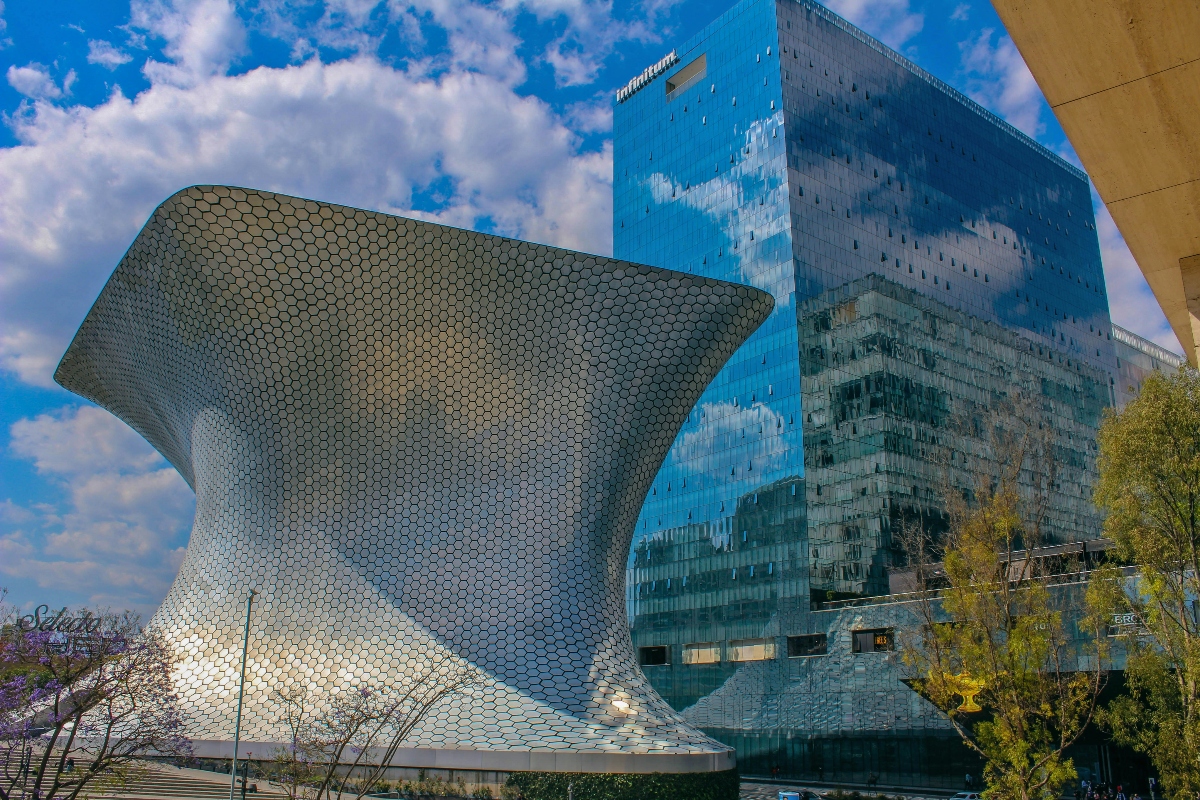 A striking contemporary building with a curved, honeycomb-patterned metallic facade stands beside a tall rectangular glass tower with a reflective blue exterior that mirrors the cloudy sky. The hexagonal pattern covers the sculptural building's organic form, while the glass high-rise features the Inbursa branding at its top. Green trees are visible in the foreground against a blue sky with white clouds.
