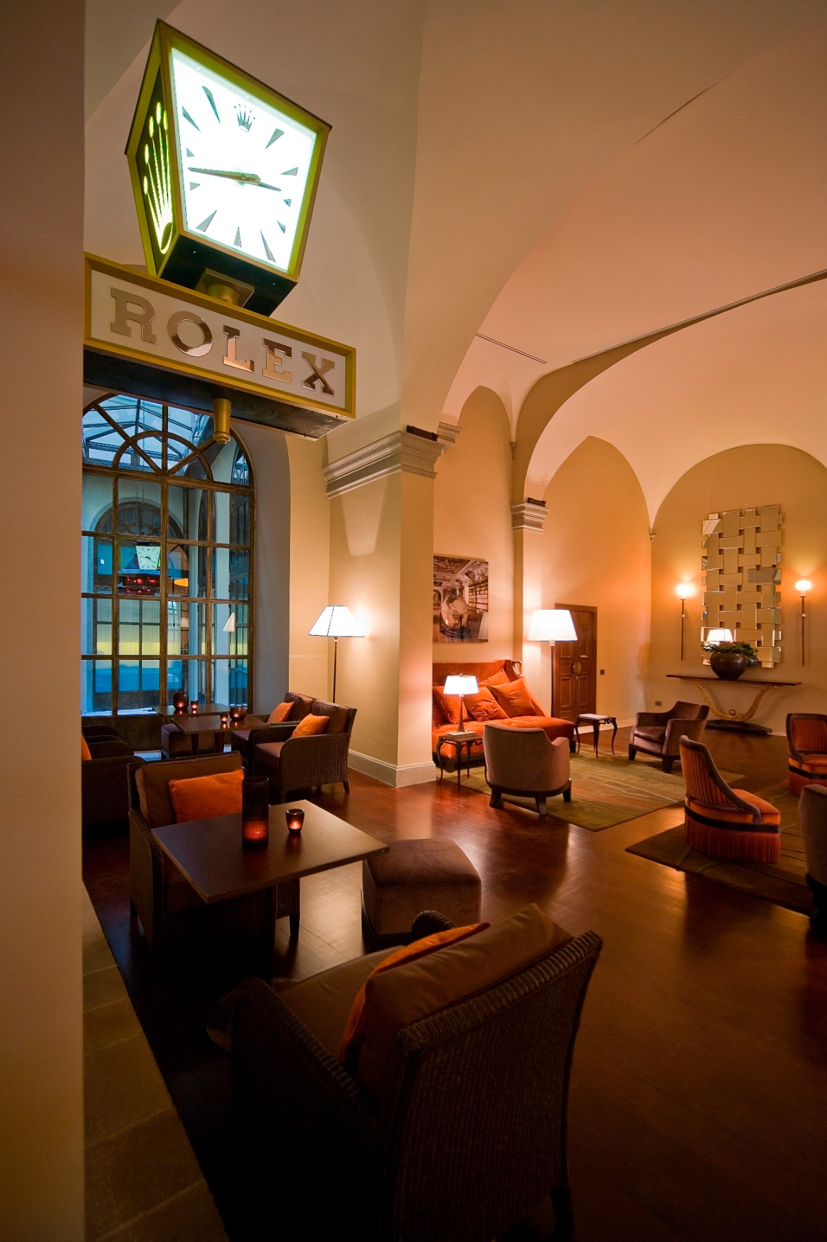 This image shows an elegant interior space featuring a prominent illuminated Rolex clock mounted on the wall above seating areas with dark upholstered furniture and orange accent pillows. The room has classical architectural elements including arched ceilings, columns, and large arched windows, with warm ambient lighting provided by table lamps and wall sconces. The space is furnished with multiple conversation areas arranged on dark polished floors, creating a sophisticated atmosphere with cream-colored walls and refined decor.