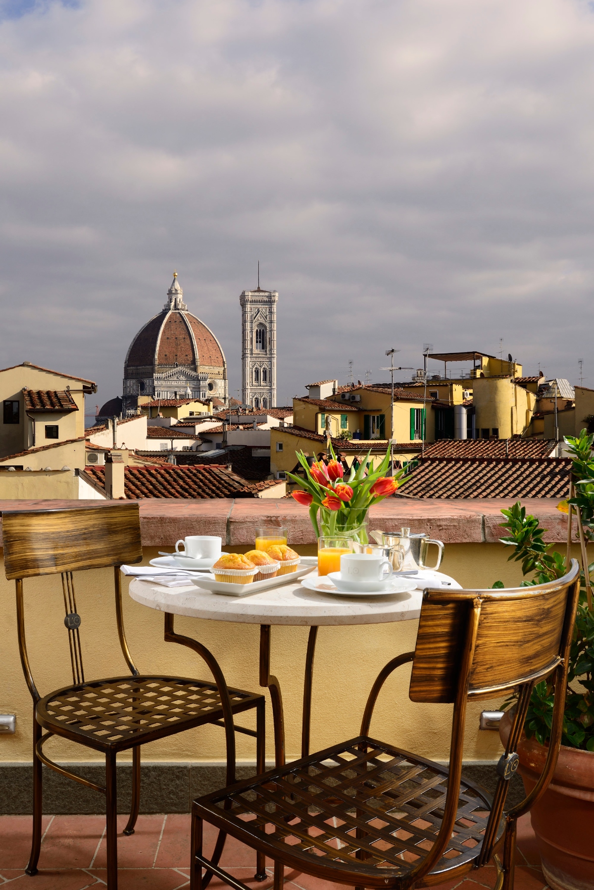 This image shows a rooftop terrace breakfast setting with a small white round table surrounded by metal chairs with latticed seats, featuring coffee cups, pastries, orange juice, and fresh flowers in a vase. The terrace overlooks a historic cityscape with distinctive architecture including a large Renaissance dome and ornate bell tower rising above terracotta-roofed buildings. The scene is set under a cloudy sky with potted plants adding greenery to the intimate outdoor dining space.
