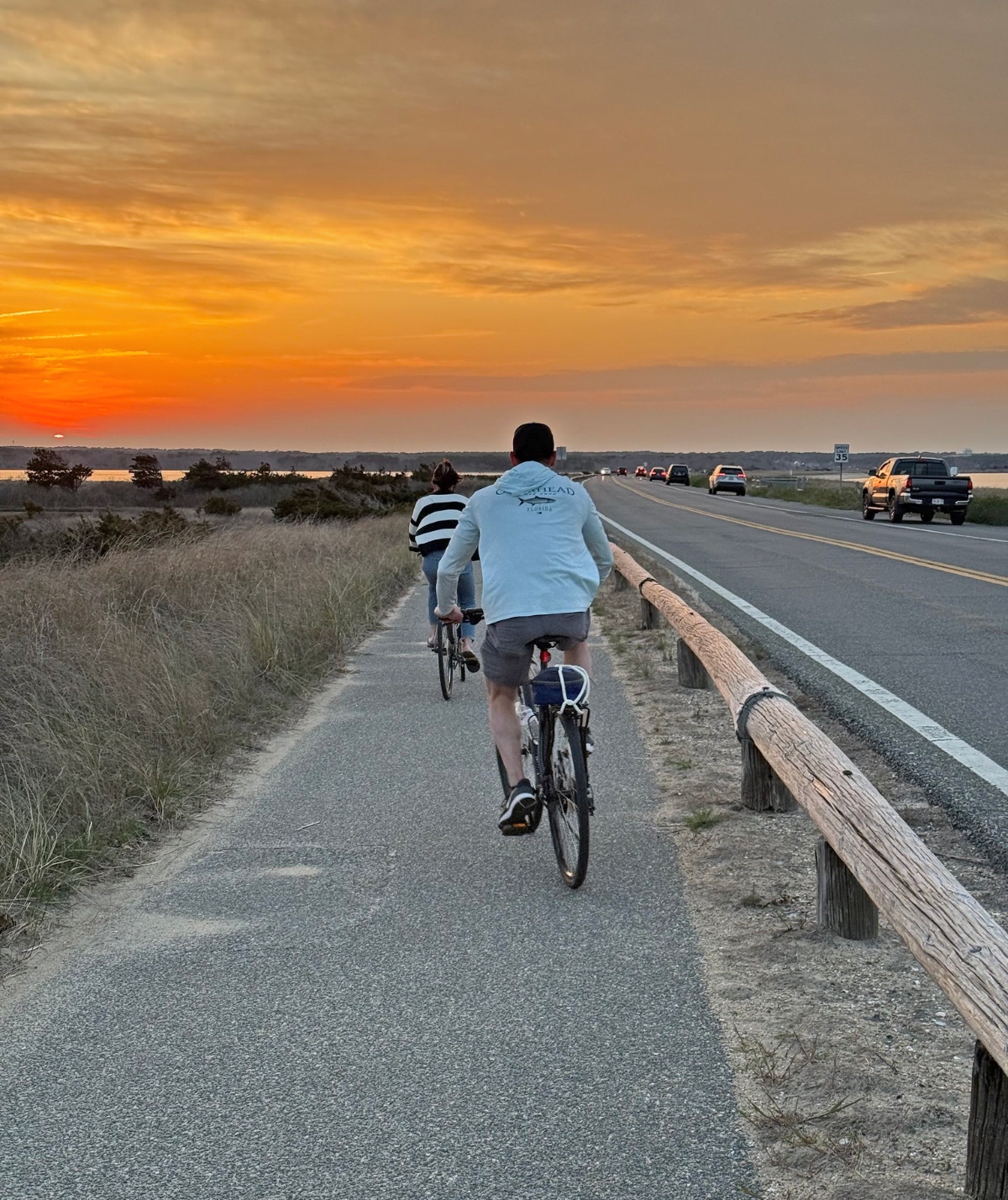 Two cyclists ride along a paved bike path that runs parallel to a highway, separated by a wooden rail barrier. The scene is set against a dramatic sunset sky with vivid orange and yellow clouds, while cars travel on the adjacent roadway through what appears to be rural grassland terrain.