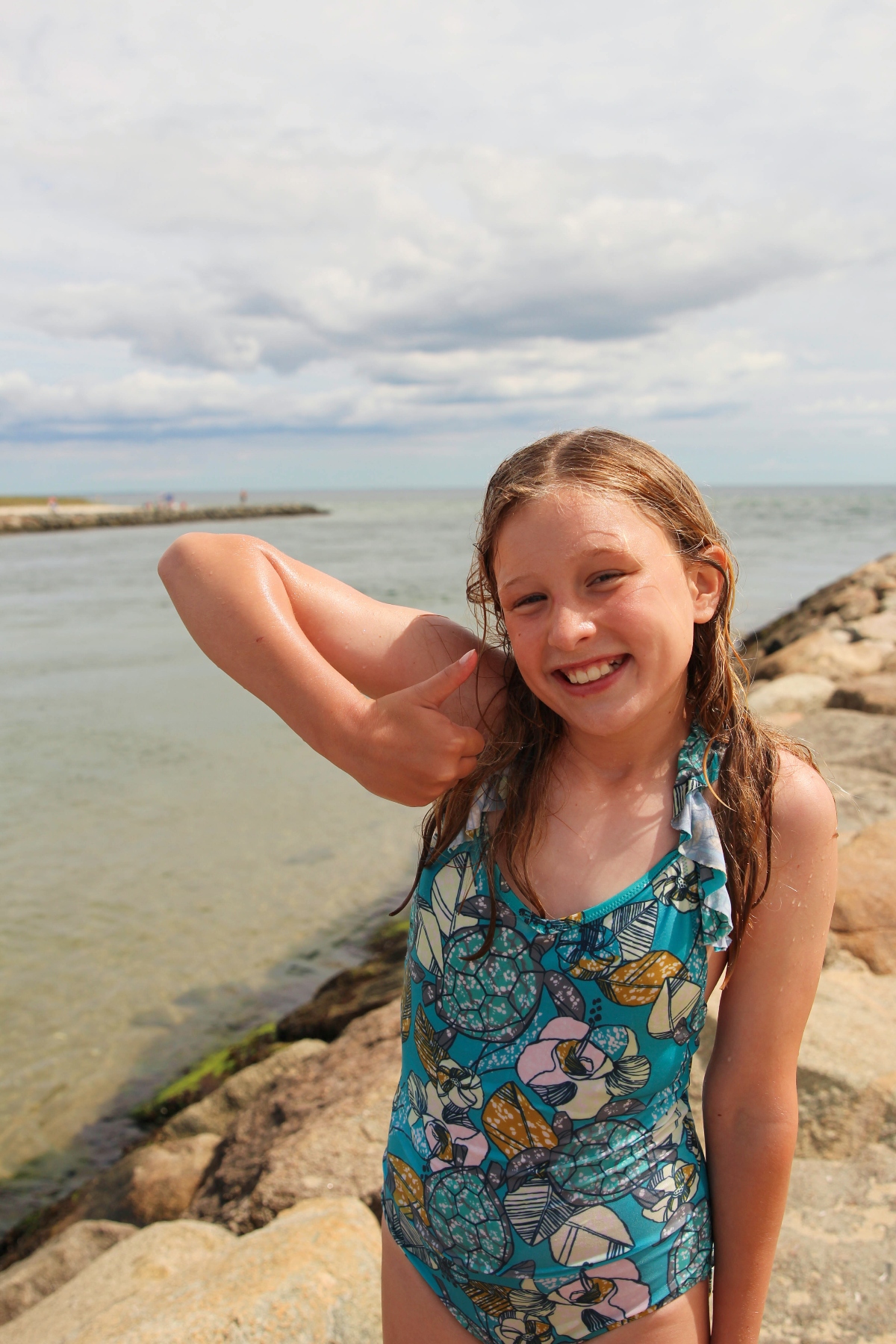 A young girl wearing a colorful patterned swimsuit stands on rocky terrain near a body of water, with her hand positioned near her head in a casual pose. The background shows calm water, a stone breakwater or jetty, and an overcast sky with gray and white clouds.