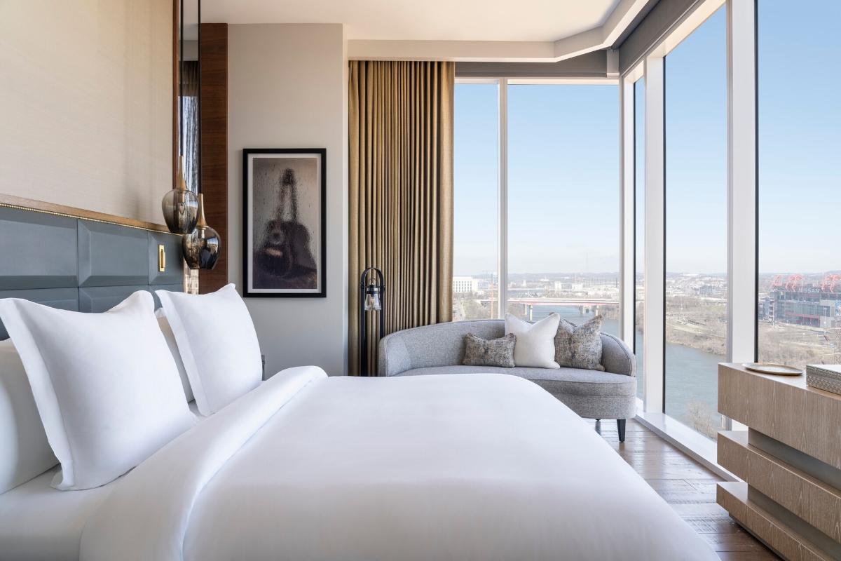 A modern hotel bedroom features a king-size bed with white linens and a gray upholstered headboard, flanked by pendant lights and decorative vases. Floor-to-ceiling windows reveal a waterfront city view, while a gray curved sofa sits near the windows beside floor-length beige curtains and minimalist wood furnishings.