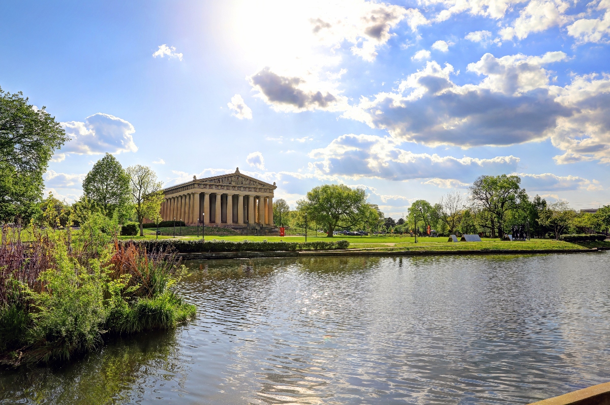 A classical columned building resembling the Parthenon sits on a grassy hill beyond a pond, surrounded by mature trees under a partly cloudy blue sky with bright sunlight. The foreground shows the pond's surface with gentle ripples and vegetation along its banks, with green lawn areas extending toward the neoclassical structure.