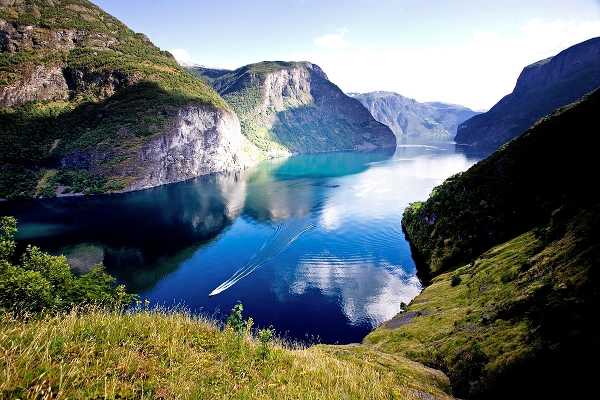 A narrow fjord with deep blue-green water winds between steep mountain cliffs covered in lush green vegetation and exposed gray rock faces. A small boat creates white wake lines as it travels through the calm water, with dramatic mountains rising on both sides and extending into the distance. Golden grass in the foreground frames the view of the fjord below.
