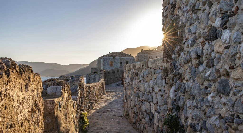 A narrow stone-paved walkway runs between weathered stone walls, with bright sunlight creating a starburst effect from behind the ancient masonry. In the background, white buildings are visible against distant mountains, while the golden lighting illuminates the rough-textured stone blocks and cobbled pathway.