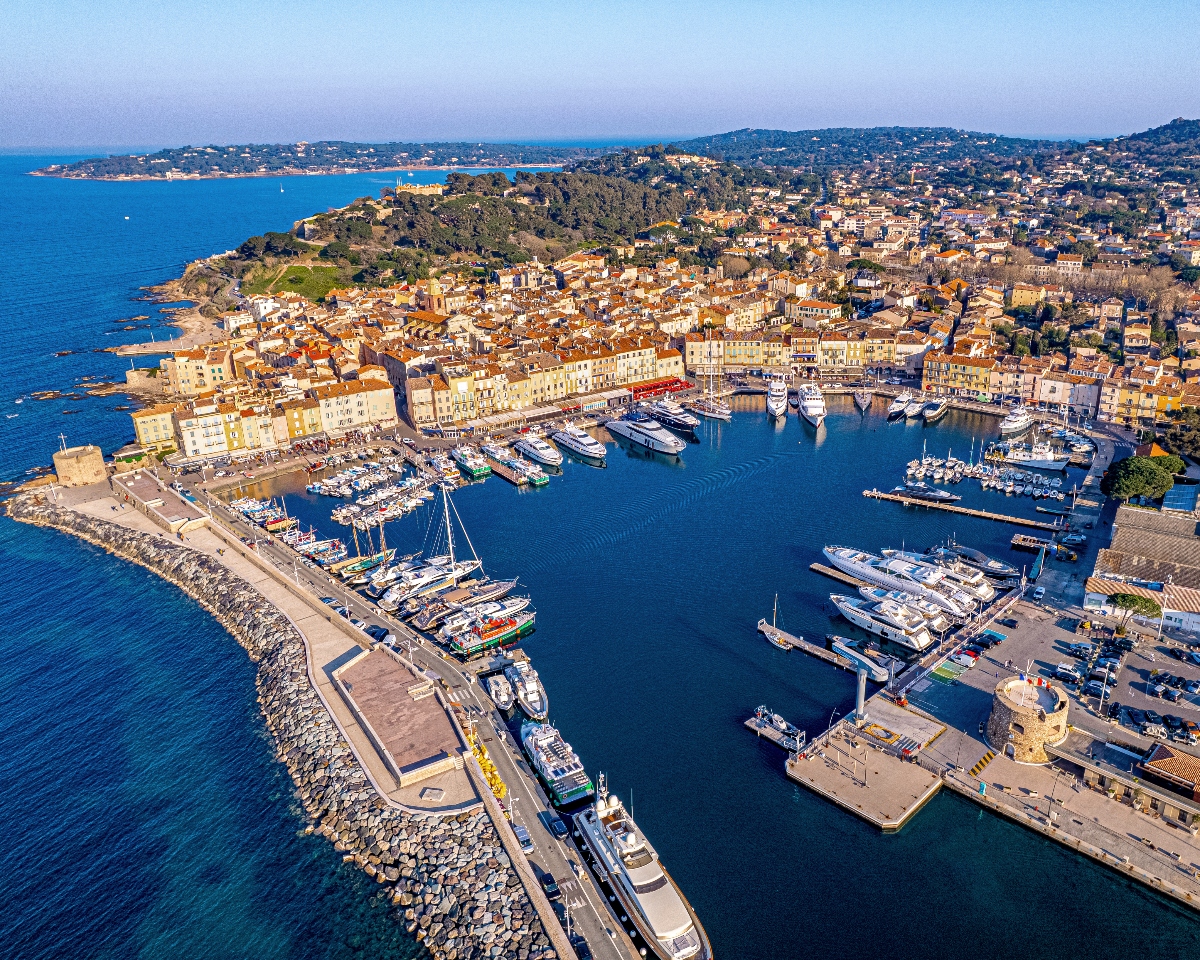 An aerial view shows a Mediterranean coastal town built around a protected harbor filled with numerous boats, yachts, and marina facilities enclosed by stone breakwaters. The waterfront is lined with colorful multi-story buildings in warm tones of yellow, orange, and cream that extend up the hillsides, creating a dense urban landscape against the backdrop of forested hills and deep blue ocean waters.