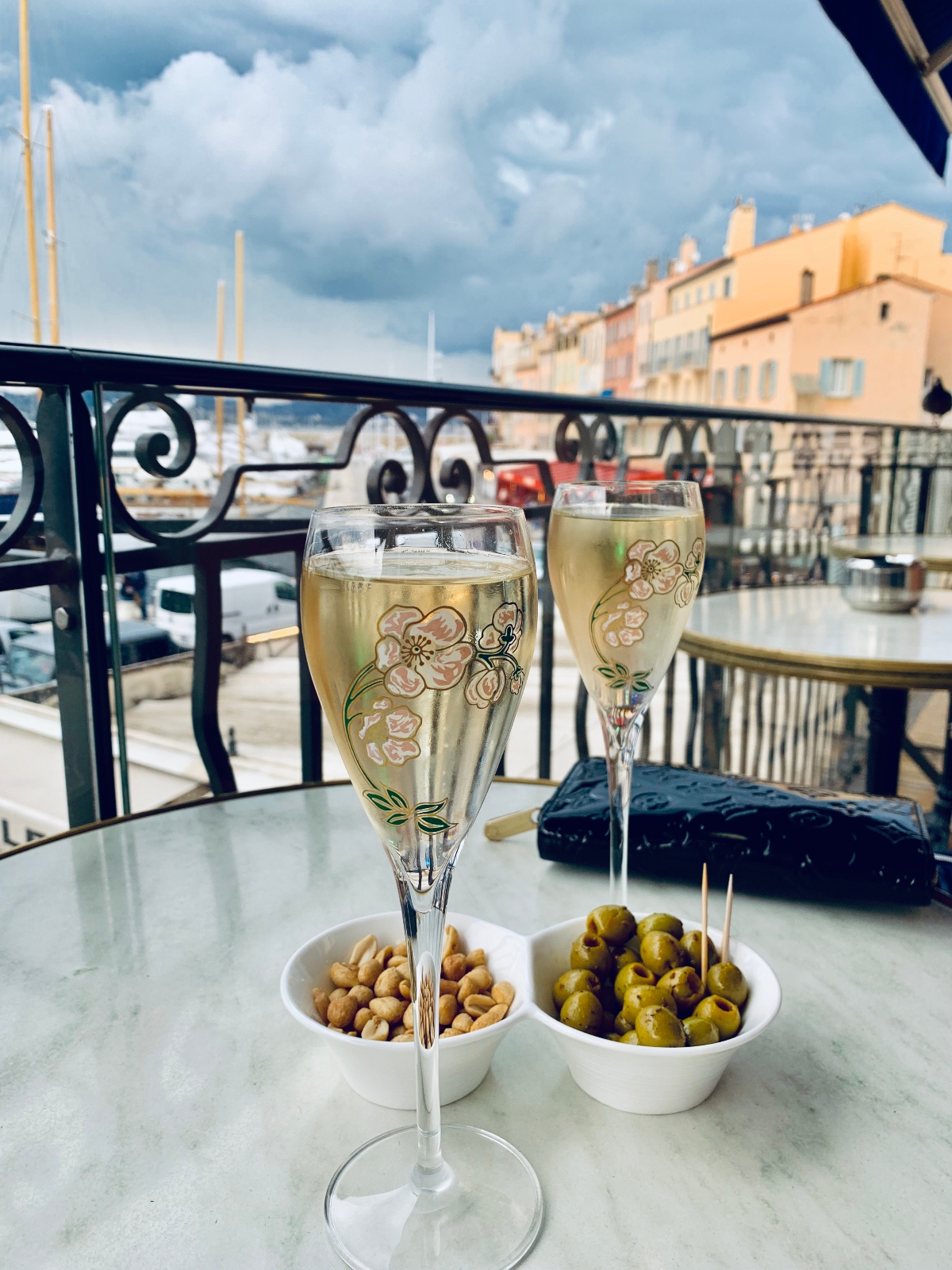 Two champagne flutes decorated with pink floral designs sit on a glass table alongside small white bowls containing nuts and green olives with toothpicks. The scene is set on a balcony with ornate black iron railings overlooking a waterfront harbor with colorful Mediterranean-style buildings and boat masts visible in the background under a cloudy sky.