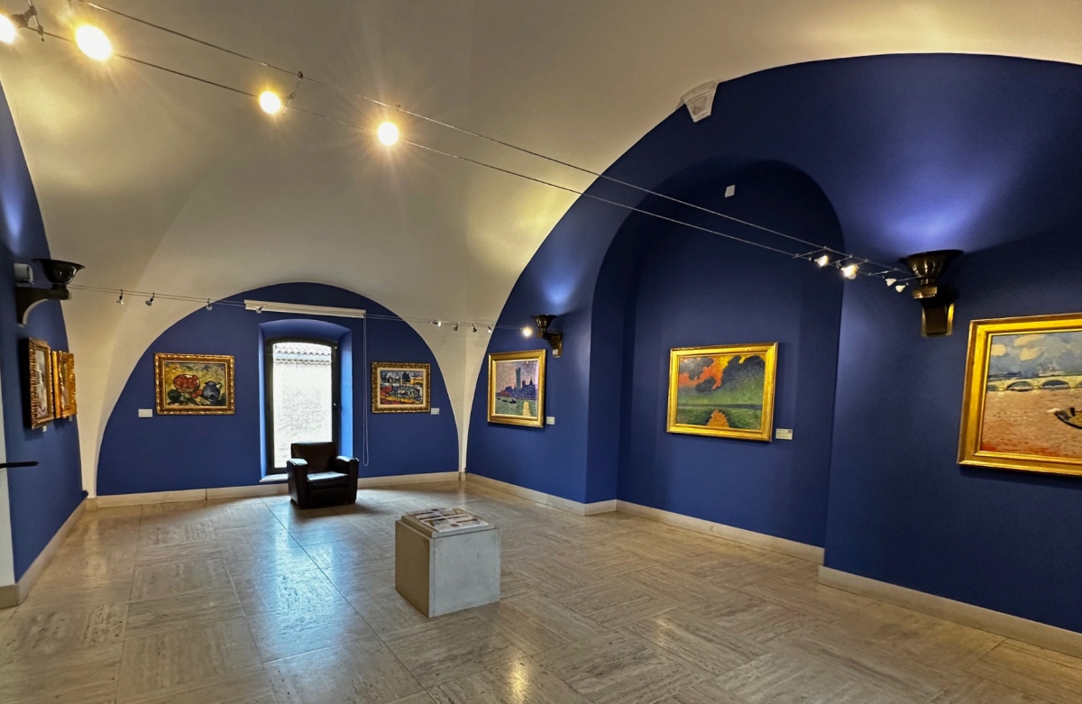 This art museum interior features deep blue arched walls and ceiling with white accent areas, displaying multiple framed paintings in ornate gold frames under track lighting. The space has a geometric patterned stone floor, a small window, dark seating furniture, and a white cubic display stand in the center of the room.