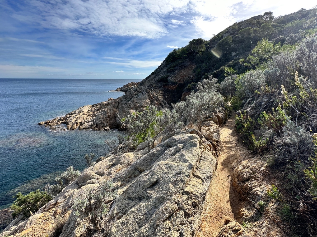 A narrow dirt hiking trail winds along a rocky Mediterranean coastline, bordered by large granite boulders and dense scrub vegetation on the hillside. The path overlooks calm blue ocean waters under a partly cloudy sky, with the rugged terrain featuring mix of exposed rock formations and native brush extending up the steep coastal slope.