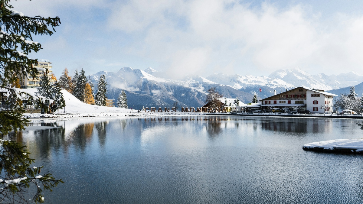 A tranquil alpine lake reflects snow-covered peaks and a cloudy sky, with large letters spelling "CRANS MONTANA" installed along the snowy shoreline. Traditional Swiss chalet-style buildings and hotels line the water's edge, framed by evergreen trees dusted with snow and some golden autumn foliage. Dramatic mountain ranges rise in the background under misty clouds, creating a scenic winter resort landscape.