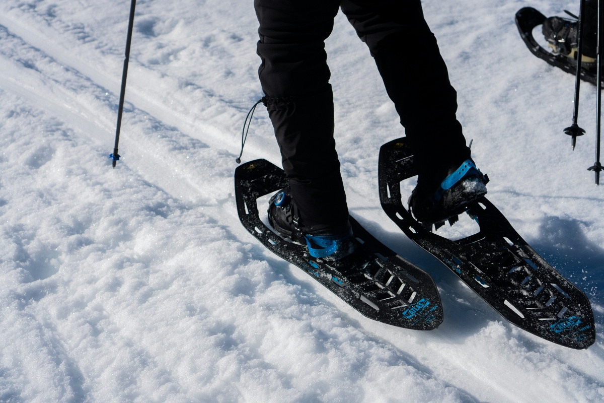 A person wearing black pants stands in black snowshoes with blue bindings and straps on packed snow. Trekking poles are visible on either side, and another snowshoe is partially visible in the upper right corner. Multiple footprints and snowshoe tracks are impressed in the white snow surface around the person's feet.