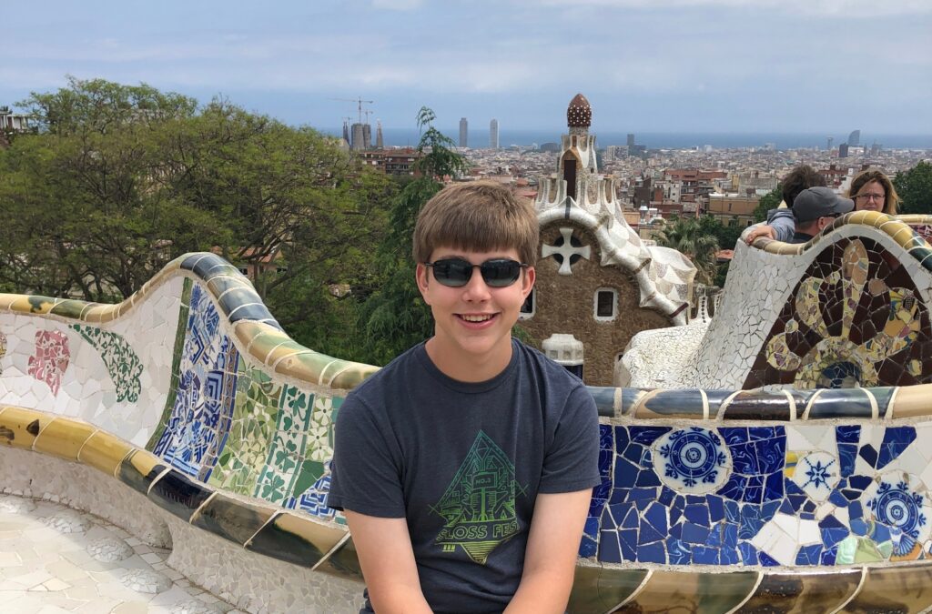 A teen boy wearing sunglasses and a dark t-shirt sits on a curved mosaic bench featuring colorful ceramic tiles in blue, green, and white patterns. The setting is an elevated viewpoint overlooking a city skyline, with trees and other visitors visible in the background.