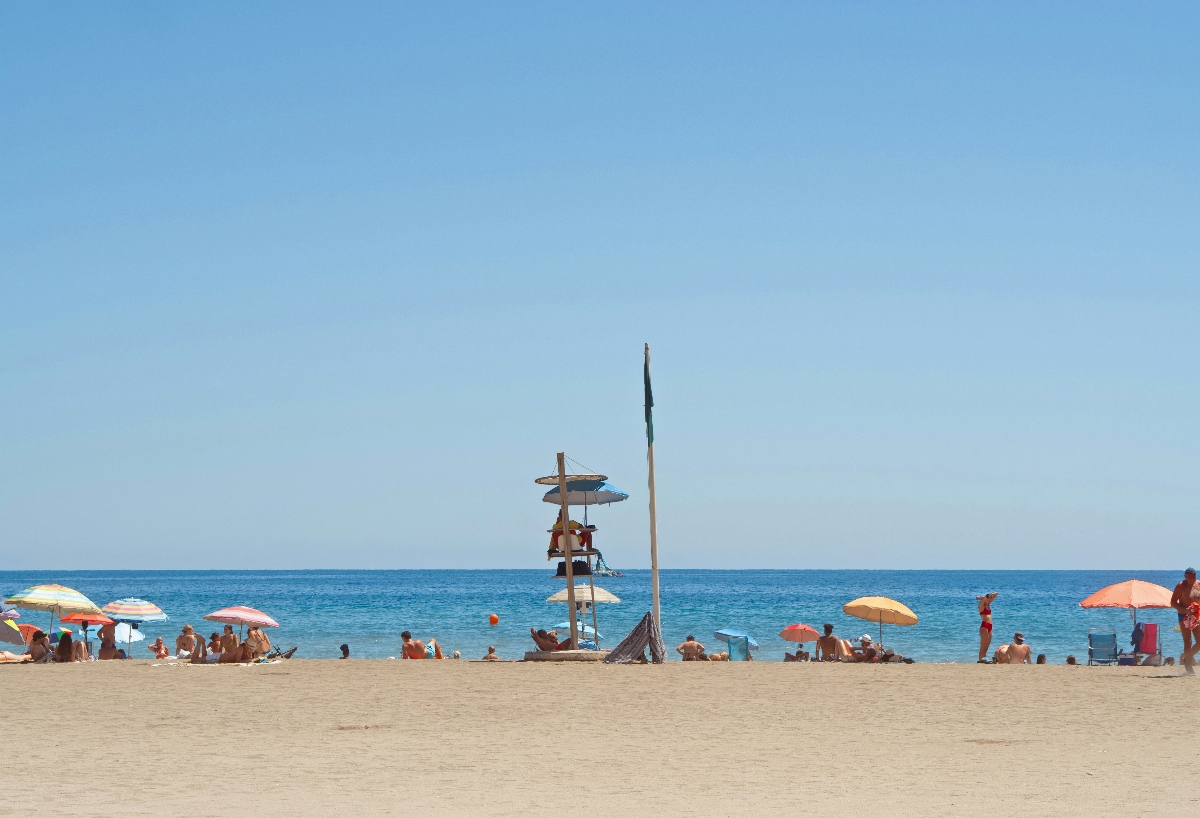 A sandy beach stretches along blue ocean water under a clear sky, with numerous beachgoers relaxing under colorful umbrellas in shades of yellow, pink, orange, and blue. A tall lifeguard station with an observation platform and flagpole stands prominently in the center of the beach scene, surrounded by people enjoying various beach activities.