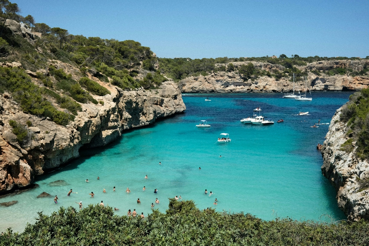 A secluded cove with brilliant turquoise and blue water is surrounded by tall limestone cliffs covered in Mediterranean vegetation. Multiple boats are anchored in the bay while numerous people swim and wade in the crystal-clear water near the rocky shoreline.